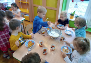 Dzieci siedzą przy stolikach i spożywają posiłek. Chłopiec samodzielnie nalewa sobie mleko z wazy.